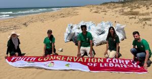 Four volunteers kneel on a sandy beach holding a fabric sign with their organization’s name (Youth Conservation Corps of Lebanon) displayed across the front.
