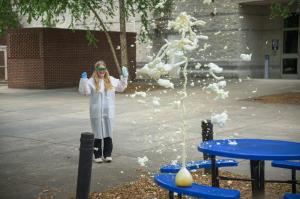 A student wearing safety goggles and a lab coat watches a foaming chemical reaction erupt from a flask during an outdoor STEM experiment at GSSM’s GoSciTech summer camp.