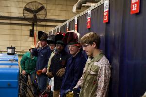 A group of young students wearing safety glasses and welding helmets stand in a row inside an industrial welding lab, leaning against blue welding booths labeled with red number signs. Large ventilation fans and workshop equipment are visible in the backg