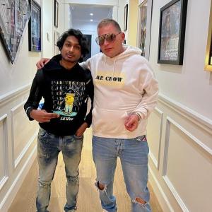Ashwin Gane and Scott Storch pose side by side in a bright hallway decorated with framed music awards and photos, both wearing hoodies and jeans.