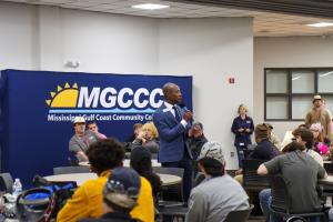 A man in a blue suit speaks into a microphone while standing in front of a Mississippi Gulf Coast Community College (MGCCC) banner, addressing a group of seated students in a classroom setting. Students sit at round tables listening, and a staff member st