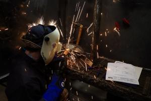 A student wearing a welding helmet, gloves, and protective gear welds metal pieces inside a welding booth. Bright sparks scatter in multiple directions, and a printed project blueprint with diagrams rests on the work surface nearby.