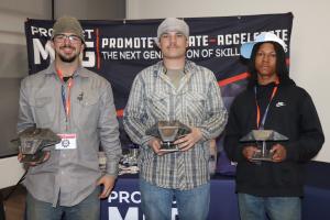 Three young men stand side by side in front of a banner reading “Promote – Educate – Accelerate the Next Generation of Skilled Trades,” each holding a metal Manta Ray unmanned underwater vehicle (UUV) project with a welded, angular body mounted on a small
