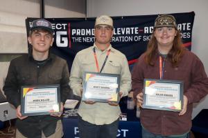 Three young men stand shoulder to shoulder in front of a “Promote – Elevate – Accelerate the Next Generation of Skilled Trades” banner, each holding a framed Certificate of Achievement. They are wearing casual work shirts, baseball caps, and event lanyard