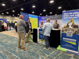Attendees talk with exhibitors at a booth on the trade show floor of the CGA Conference & Expo. Several people wearing conference badges stand around a display for the Damage Prevention Action Center, with banners, a QR code sign, and a hashtag sign readi