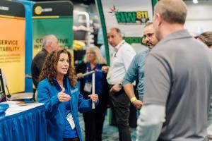 A woman wearing a blue jacket speaks with two attendees at an exhibit booth during the CGA Conference & Expo, gesturing with her hands while holding a white piece of equipment. Other attendees and booth displays, including signage for 811 damage preventio
