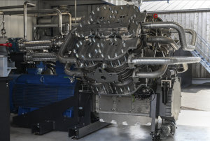 The photo shows an industrial heat pump which looks like a big truck engine made out of stainless steel. It has many bolts on it and it is resting on metal springs to absorb any vibrations. There are big pipes going in and out of the machine which supply 