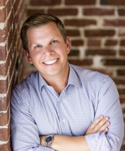 Smiling man in a checkered shirt leans against a brick wall, arms crossed. The setting is bright, conveying a friendly and approachable mood.