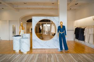 Interior of the Jendue flagship store at Town Center Boca Raton, featuring gold mannequins styled in denim and white apparel, marble cylindrical display pedestals, a circular illuminated clothing rack set against a marble wall panel, and curated garment r