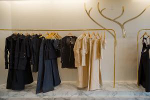 Interior clothing display at the Jendue flagship store at Town Center Boca Raton, featuring gold rail garment racks lined with limited-edition pieces including dark navy denim long coats, wide-leg jeans, dark zip-up jackets, and beige trench-style coats w