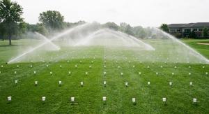 Rows of white catch cans spread across a green commercial lawn with rotor sprinklers spraying water during an irrigation audit test