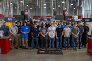 A group of students and instructors pose together inside a modern manufacturing training facility, standing in front of CNC machines and banners for Project MFG. The group is arranged in several rows on the shop floor, wearing casual clothing and work att
