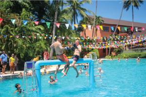 A lively pool scene with individuals competing on an inflatable platform, surrounded by colorful flags, palm trees, and a tropical resort backdrop.