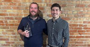 Christopher Gayler holding an Apex Operations award standing next to a colleague against a brick wall background during a team celebration.