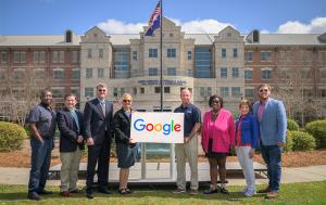 A group of nine adults stand outdoors in front of a large academic building with American and state flags. Two people in the center hold a sign with the Google logo while the others stand on either side, posing for a photo on a lawn with the building entr