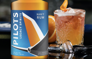 Pilot’s Legacy Navy Strength Rum bottle beside a rum cocktail with orange garnish and ice in a crystal glass.