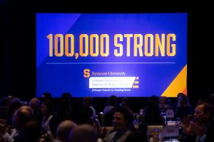 Screen centered in image with the words 100,000 Strong. People seated in dark room in front of the screen.