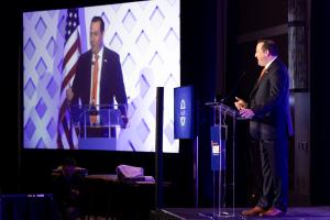 Presenter stands at podium with a screen next to the stage showing the presenter's face. 