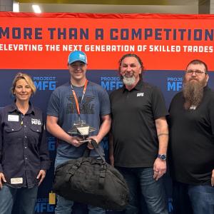 Four people stand in front of a orange and blue Project MFG banner that reads “More Than a Competition: Elevating the Next Generation of Skilled Trades.” The person in the center holds a welded model mantra ray UUV and a large duffel bag while the others 