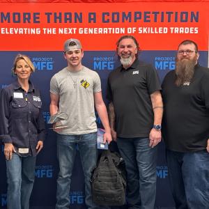 Four people stand in front of a orange and blue Project MFG banner that reads “More Than a Competition: Elevating the Next Generation of Skilled Trades.” The person in the center holds a welded model mantra ray UUV and a large duffel bag while the others 
