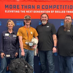 Four people stand in front of a orange and blue Project MFG banner that reads “More Than a Competition: Elevating the Next Generation of Skilled Trades.” The person in the center holds a welded model mantra ray UUV and a large duffel bag while the others 