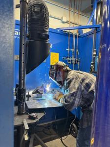 A person wearing a welding helmet and protective gloves welds a metal piece on a workbench inside a blue welding booth. Bright sparks are visible where the welding torch meets the metal, and ventilation equipment and pipes are mounted above the workstatio