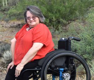Pamela wearing a red blouse. black pants, and glasses, leans forward as she sits in her wheelchair.  The setting is outdoors and in the desert of Tucson. She is smiling and engaged with the viewer. 
