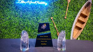 Crystal awards trophies for GlowRow on a black table, featuring Innovative Experience of the Year (Texas/USA) and others, against a green foliage wall with "I Like A Good Paddling" neon sign, wooden paddles, and canoe backdrop.
