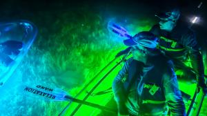 Two people in a transparent kayak with green LED glow, one holding a paddle labeled "RELAXATION WAND," surrounded by illuminated water at night.