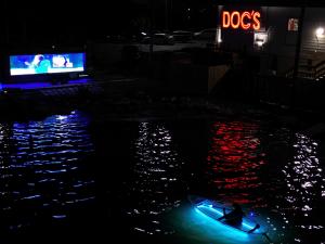 Nighttime photo of a large illuminated trailer screen playing a movie, reflecting on water with red and blue hues, a single blue-glowing kayak with paddler in foreground, and red neon "DOC'S" sign on building in background.