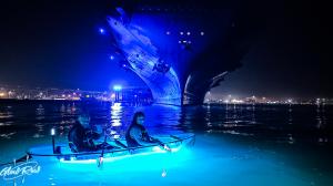 Two people in a transparent blue-lit kayak paddling toward a large blue-illuminated ship hull at night, with city lights in the background and water reflections, featuring GlowRow logo.