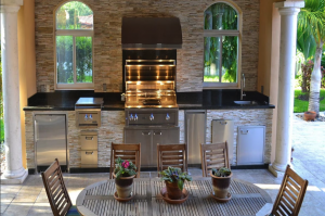 Luxury outdoor kitchen with natural stone backsplash, black granite countertops, built-in grill with hood, and stainless steel appliances under a covered patio