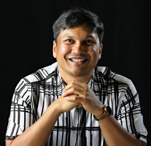 A smiling man in a plaid shirt, seated with his hands under his chin.