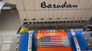 A Barudan embroidery machine stitching an American flag patch onto a bright orange safety vest.