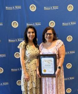 Assemblywoman Blanca E. Rubio and Maryvale Chief Program Officer Christina Moore stand together smiling in front of a floral backdrop displaying the Assemblywoman Blanca E. Rubio District 48 Women of the Year Ceremony banner. Christina Moore holds a frame