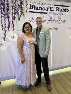 Maryvale Chief Program Officer Christina Moore and President and CEO Steve Gunther stand together smiling in front of a floral backdrop displaying the Assemblywoman Blanca E. Rubio District 48 Women of the Year Ceremony banner. Gunther attended in support