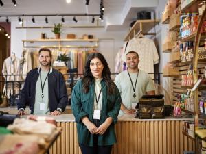 Three retail store employees wearing lanyards stand together behind a checkout counter, smiling at the camera. Clothing racks and product shelves are visible in the background.