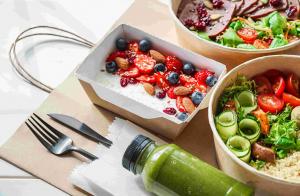 Selection of ready to eat foods and salads in takeaway packaging alongside bottle of smoothie and knife and fork.an