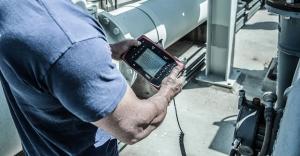 A technician in a blue shirt uses a handheld digital vibration analyzer to monitor industrial machinery. The device screen displays a real-time waveform graph, illustrating the diagnostic and measurement phase of an acoustic engineering project.