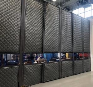 Industrial floor-to-ceiling acoustic curtain system with a diamond-quilted grey texture and clear horizontal vision panels. The sound-blocking barrier is suspended in a warehouse or factory setting to isolate noisy machinery.