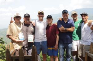 A group of seven men and teenagers stand in a row, smiling and embracing against a breathtaking backdrop of lush, rolling green mountains under a bright sky. They are dressed casually in t-shirts and baseball caps, with several wearing Sur Coffee branded clothes.