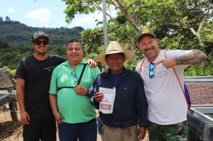 Four men stand together smiling at a coffee farm in El Salvador under a bright sky. A local producer in a straw hat holds a white bag of Sur Coffee "El Salvador" beans, while Sur Coffee team members in casual attire and baseball caps stand beside him. In
