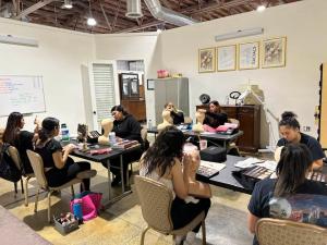A classroom of cosmetology students working at tables, practicing makeup application and facial techniques on mannequin heads. The room features professional lighting, makeup kits, and a whiteboard with lesson notes