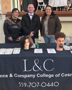 Three representatives from Lawrence & Company College of Cosmetology standing behind a promotional table. The table features a black "L&C" branded tablecloth, informational brochures, and two mannequin heads showcasing hair and beard styling.