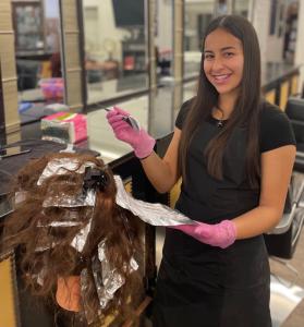 A smiling cosmetology student wearing pink gloves and a black apron practices hair highlighting. She is applying color with a brush onto hair foils on a mannequin head in a salon classroom setting