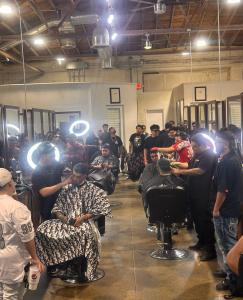 A busy, high-energy barbering classroom filled with students. Barbers are performing precision fades and hair designs on clients in professional chairs under bright ring lights, creating a modern shop atmosphere