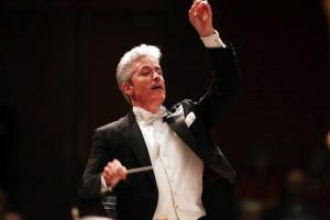 Photo of conductor Steven Byess at the podium, holding a baton in his right had and his left hand is raised up. He is wearing a black  tuxedo with white bow tie.