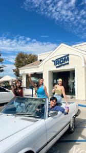 Four friends smiling and holding iced beverages while posing in a white convertible parked in front of a Tram Cream Coffee storefront on a bright, sunny day.