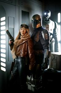 Two patrons pose in front of a K2SO droid. The person on the left wears a simple leather jacket, scarf and goggles, while the person on the right is dressed in a full Mandalorian style armor set. The background is inspired by Star Wars imperial architectu