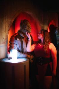 A guest dressed as a lizard-like Trandoshan alien looks down at a human woman. In the backround are familiar arched alcoves and stucco walls inspired by the Mos Eisley cantina from Star Wars